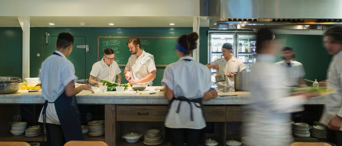 large kitchen prep area with cooks, servers, and restaurant equipment in background